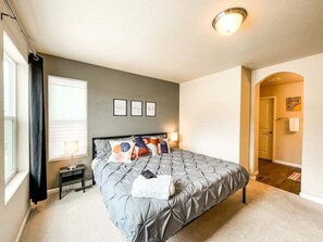 A bedroom with a bed, bedside tables, a window with curtains, a wall with framed pictures, and a hallway. - 2102 Birmingham (Colorado Springs)
