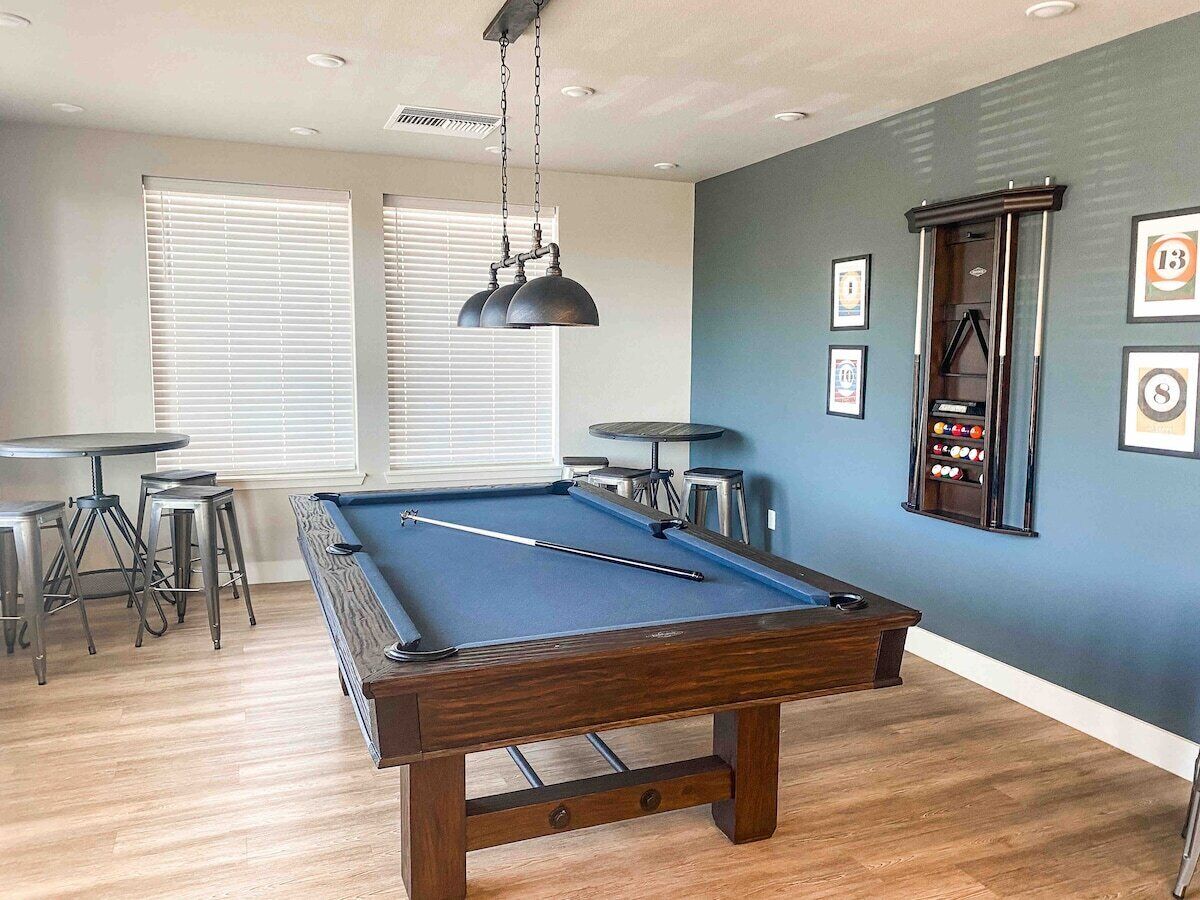 A pool table in a room with blue walls, wooden flooring, and a ceiling fan. There are framed pictures on the wall and a pool table with stools around it.