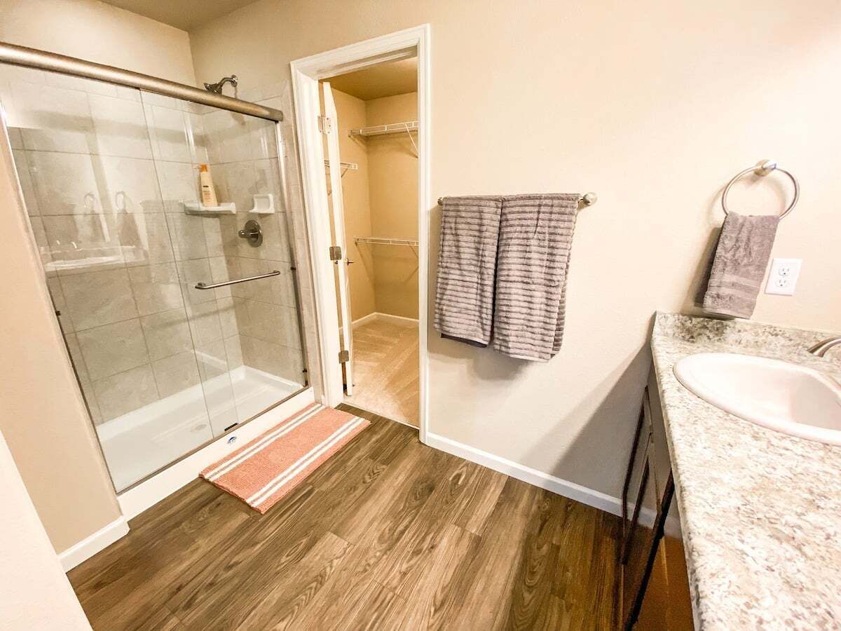 Une salle de bain dotée d’une douche à parois de verre, d’un plancher en bois, d’un lavabo avec un comptoir en granit et d’un porte-serviettes avec des serviettes.