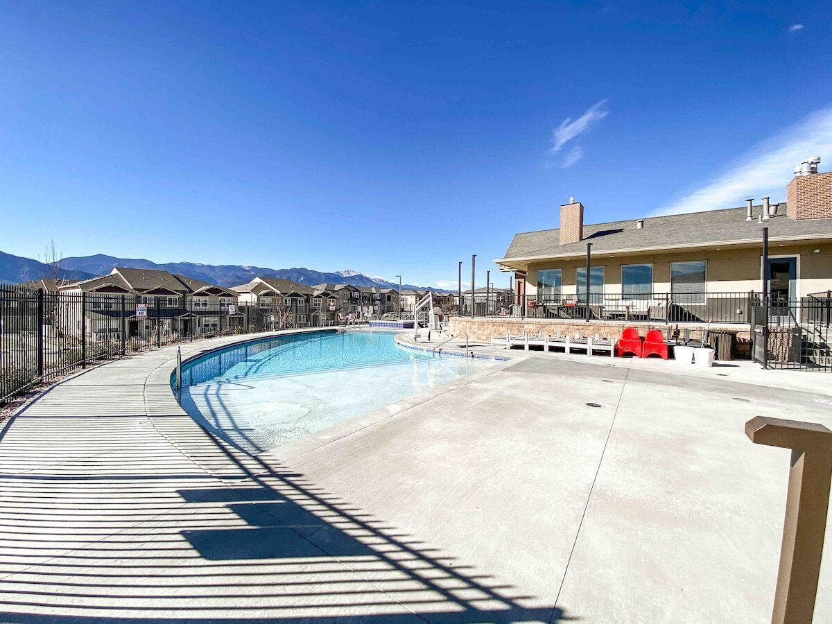 A swimming pool with a curved deck, surrounded by a fence, and a building with a chimney in the background.