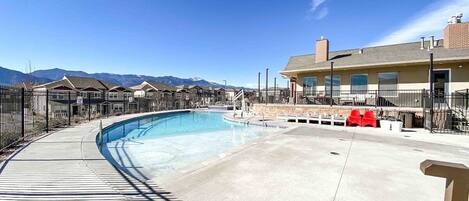 A swimming pool with a curved deck, surrounded by a fence, and a building with a chimney in the background.