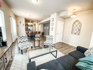 A modern living room with a sofa, two armchairs, a glass coffee table, and a dining area in the background. - 2102 Birmingham (Colorado Springs)