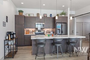 A modern kitchen with dark cabinets, a central island with bar stools, stainless steel appliances, and pendant lighting. - Rsd 3040 (Coeur d'Alene)