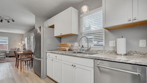 A modern kitchen with white cabinetry, a stainless steel refrigerator, a double sink, and a wooden dining table with chairs. - 1313 Cooper (Colorado Springs)
