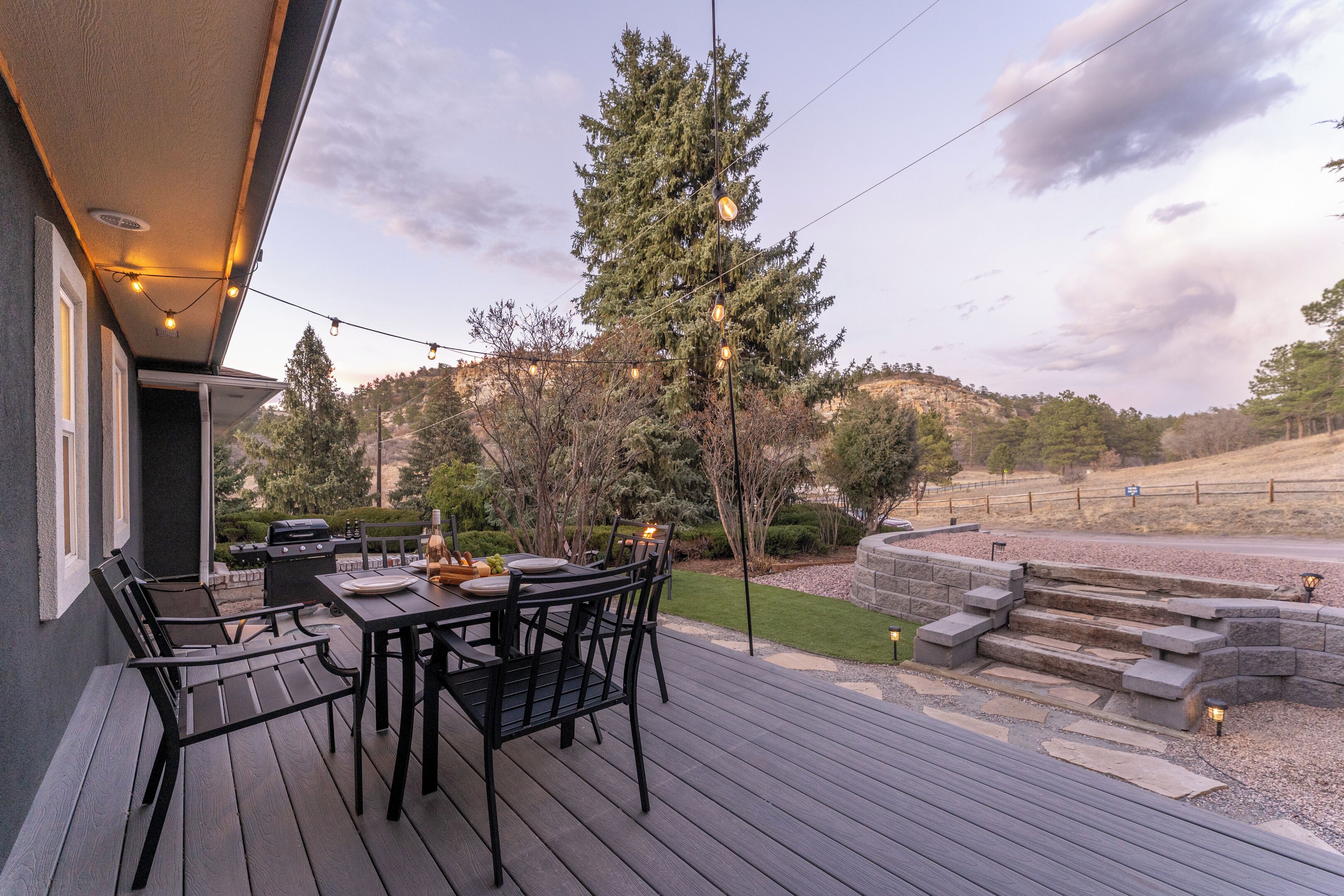 An outdoor deck with a table and chairs, surrounded by a landscaped garden and a view of a hill.