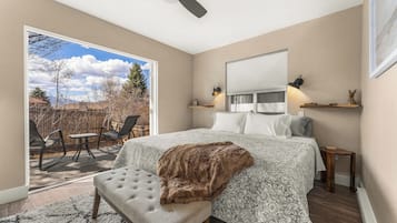 A bedroom with a large bed, a ceiling fan, a window with blinds, and a view of the backyard.