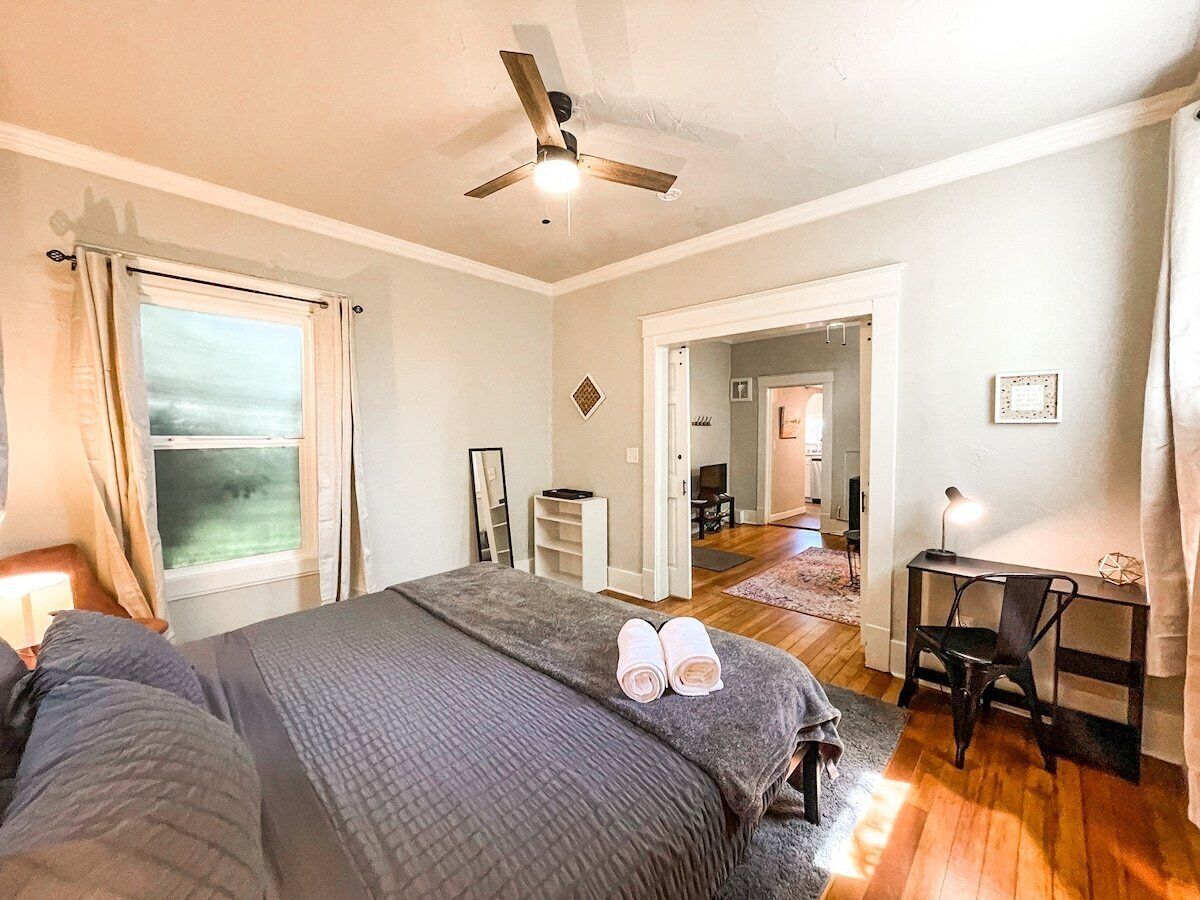 A bedroom with a bed, a desk, and a chair. There is a window with curtains, a ceiling fan, and a door leading to another room.