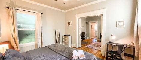 A bedroom with a bed, a desk, and a chair. There is a window with curtains, a ceiling fan, and a door leading to another room.