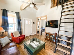 A living room with a red armchair, a glass coffee table, a wooden bookshelf, a TV mounted on the wall, and a ceiling fan. - 1208 W Colorado (Colorado Springs)