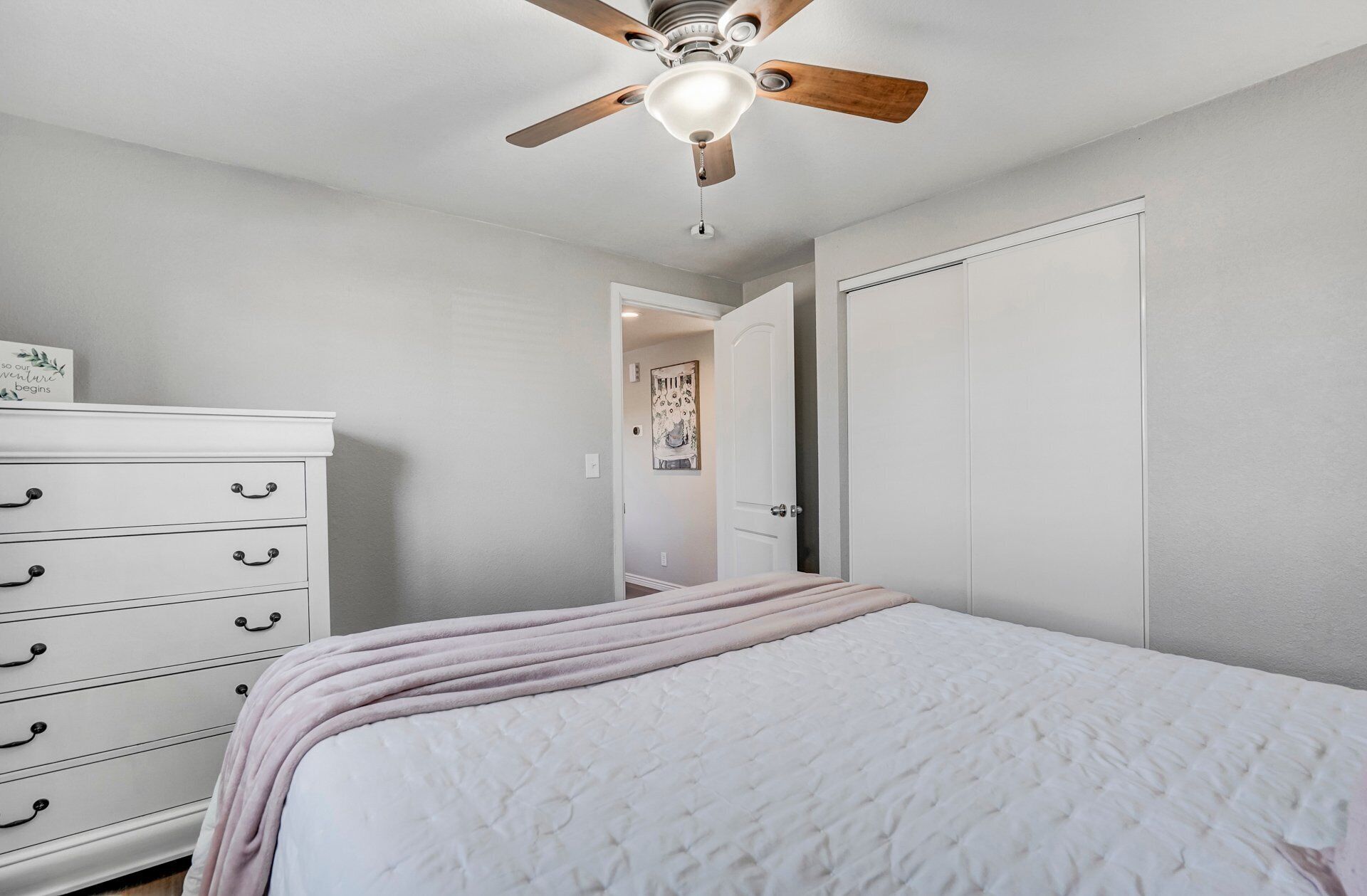 Un dormitorio con cama, cómoda, ventilador de techo y una puerta que da a otra habitación.