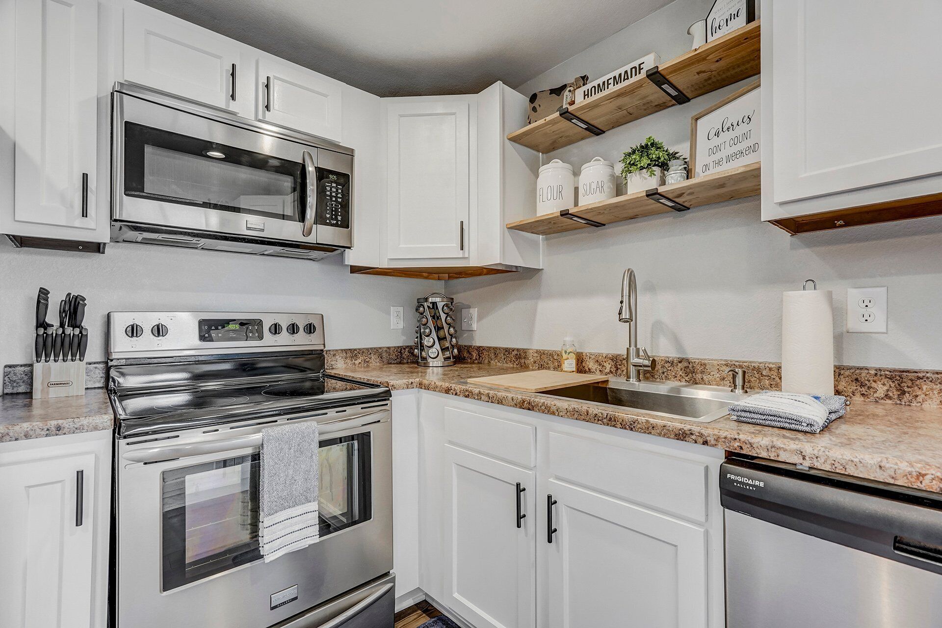 Una cocina moderna con armarios blancos, electrodomésticos de acero inoxidable y repisas de madera con utensilios de cocina.