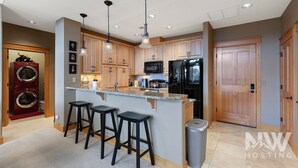 A modern kitchen with wooden cabinets, a black refrigerator, a microwave, and a built-in oven. There are three black bar stools and a trash can. - RC 2050 309 (Coeur d'Alene)