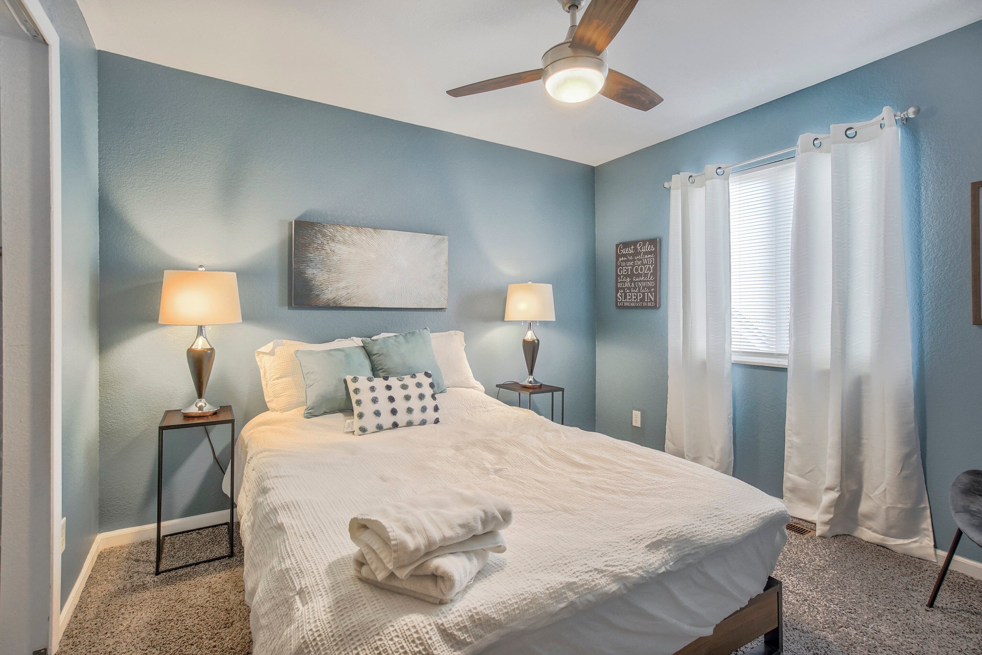 A bedroom with a bed, two bedside lamps, a ceiling fan, a window with curtains, and a wall art piece.