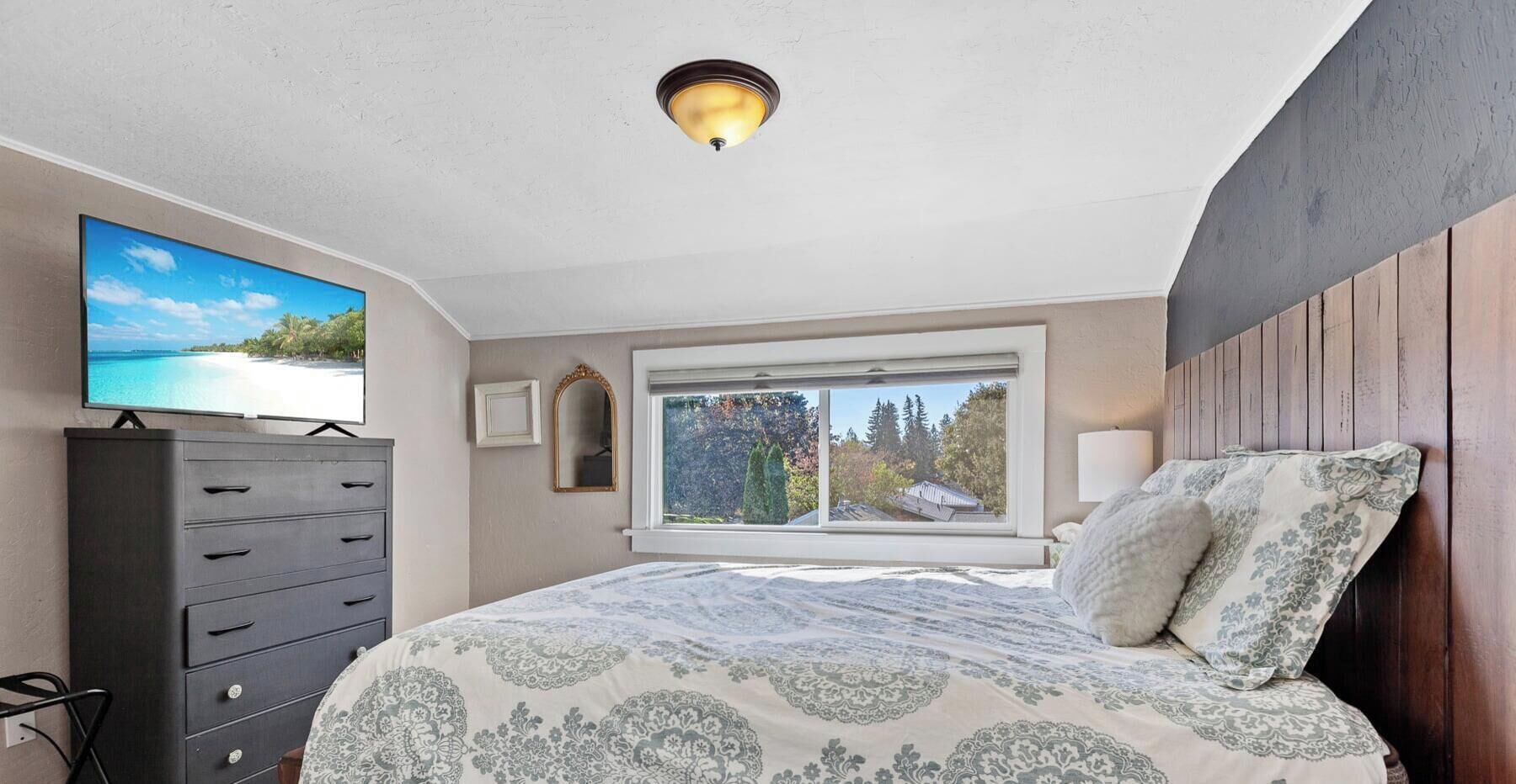 A bedroom with a bed, dresser, TV, window, and ceiling light.