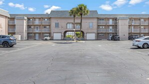 Un edificio de apartamentos de varios pisos con entrada central, espacios de estacionamiento y una palmera.