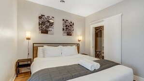 A neatly made bed with a grey quilt, two wall-mounted lamps, and a door leading to a closet. - Downtown Getaway With Balcony King Beds (Colorado Springs)