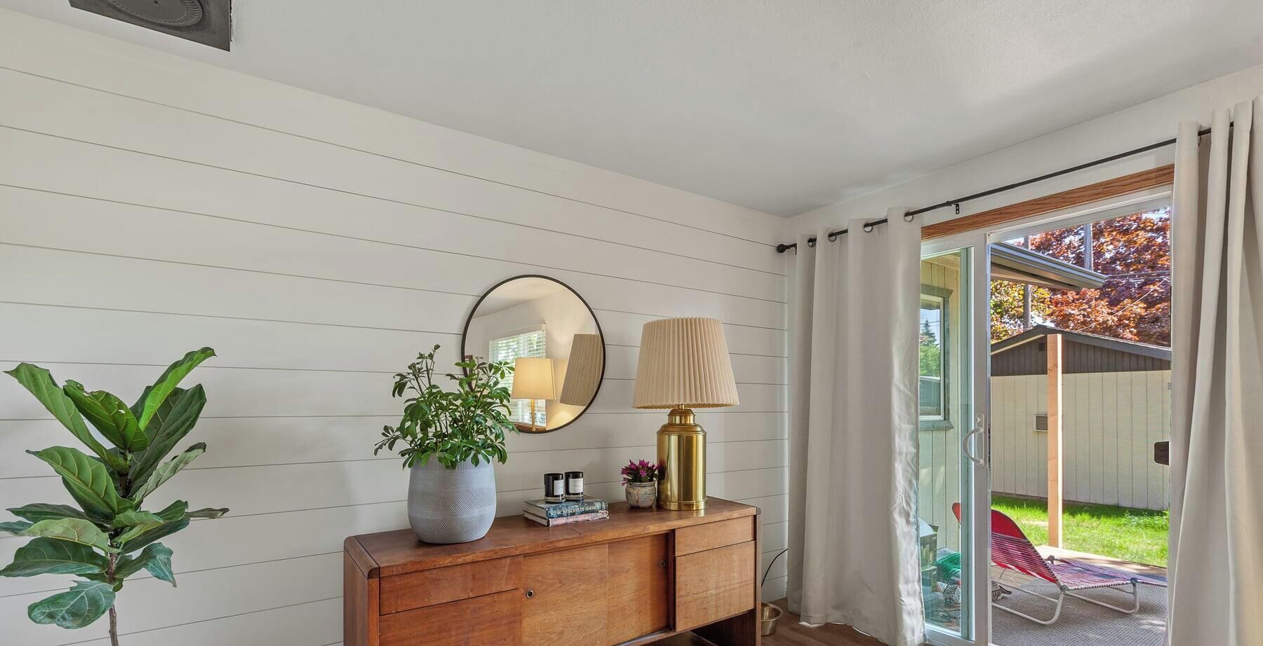 Una habitación con un aparador de madera, un espejo, una planta en maceta, una lámpara y una puerta corrediza de vidrio que da a un área exterior.