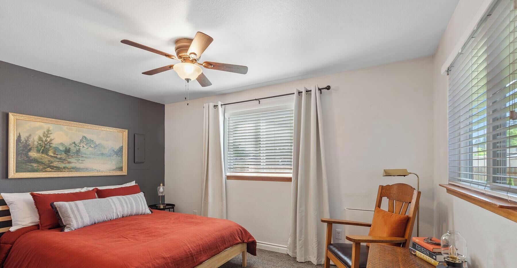 A bedroom with a bed, a chair, a ceiling fan, a window with blinds, and a painting on the wall.