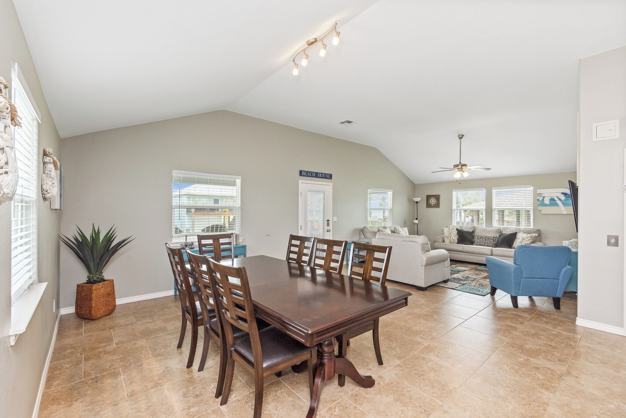A spacious dining area with a large wooden table and chairs, a ceiling fan, and a view of the living room with a sofa and armchairs.