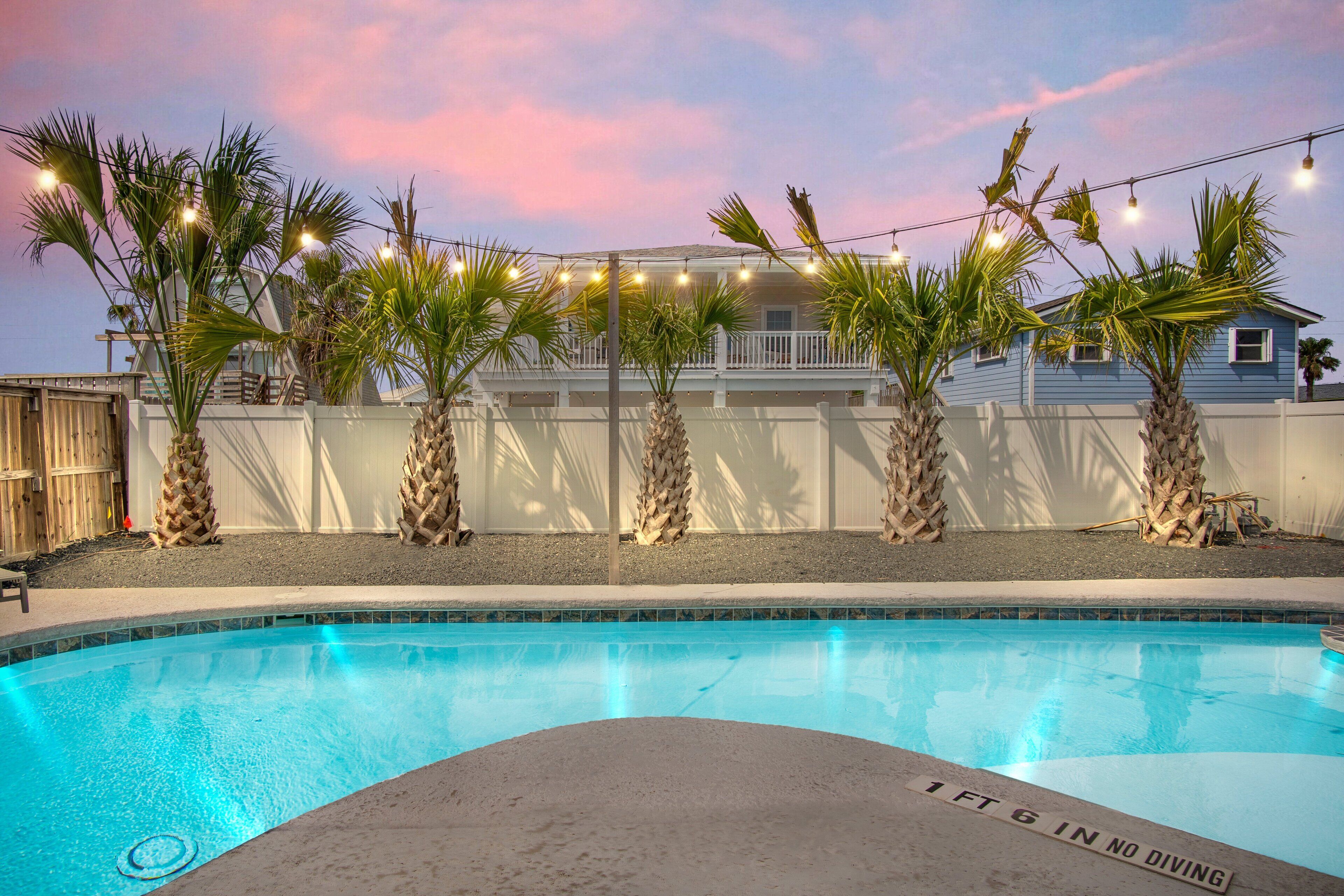 A swimming pool with palm trees and string lights, surrounded by a white fence and a building in the background.
