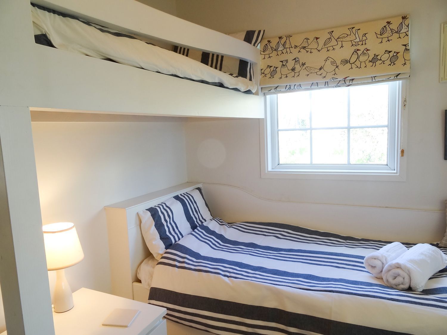 Una cama individual con ropa de cama a rayas, una lámpara de noche y una ventana con vistas a la vegetación.