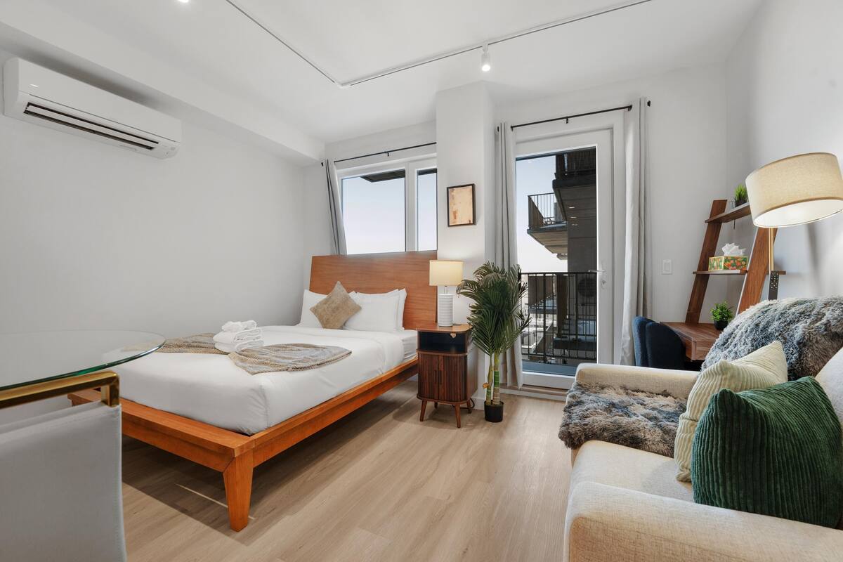 A modern hotel room with a wooden bed, a glass table, a sofa, a lamp, and a view of the outside balcony.