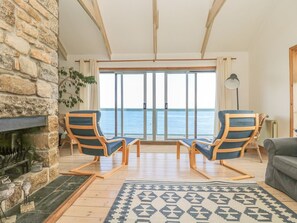A cozy living room with a stone wall, two wooden-framed chairs, a fireplace, and a view of the sea through large glass doors. - Harbour Lights (Penzance)