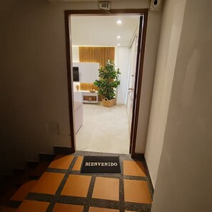 Ein Hotel Eingang mit einer Fußmatte mit der Aufschrift 'BIENVENIDO', einem Blumentopf und einem Empfangsbereich inklusive Fernseher und dekorativer Wandverkleidung.