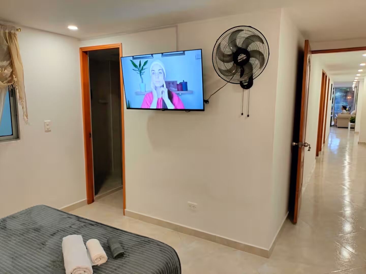 A hotel room with a bed, a TV mounted on the wall, a standing fan, and a door leading to another room.