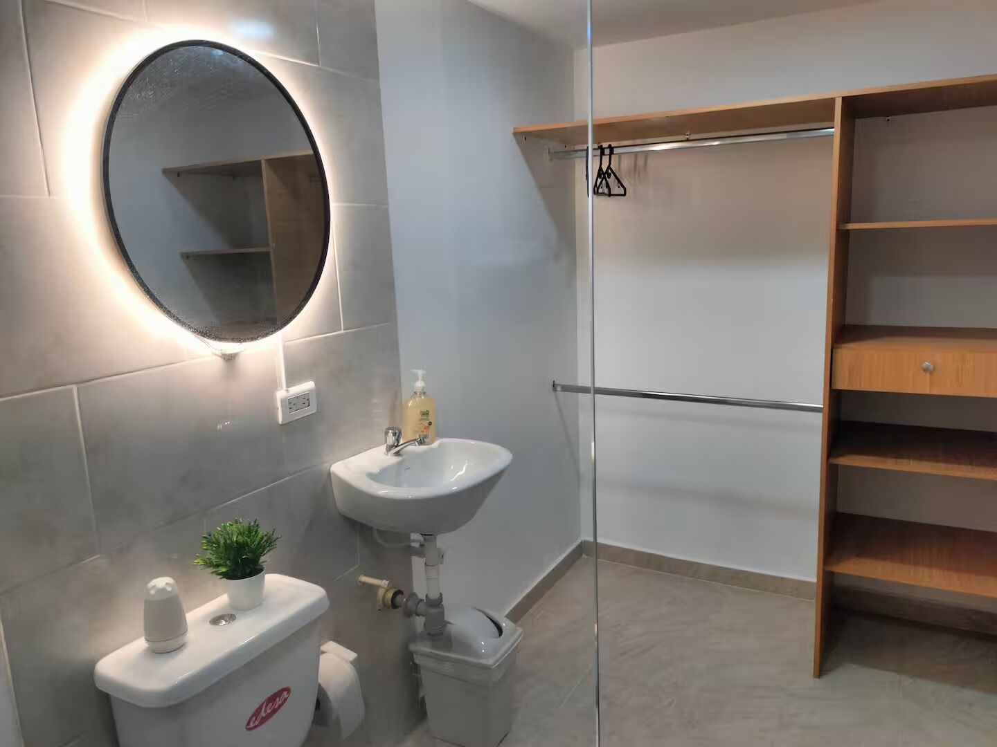 Un baño moderno con inodoro, lavamanos y espejo. Hay una planta pequeña sobre el inodoro, un dispensador de jabón en el lavamanos y una toalla colgada en una barra de metal.
