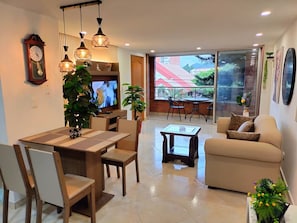 A modern living room with a dining area, a sofa, and a TV. There is a large window with a view of buildings and trees. - Exclusive, luxurious, centrally located apartment — serene and comfortable 302 (Medellín)