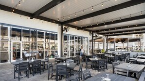 An outdoor dining area with tables and chairs, large windows, and a covered section with string lights.