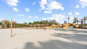 A sandy beach volleyball court with a net, palm trees, and a water feature in the background.
