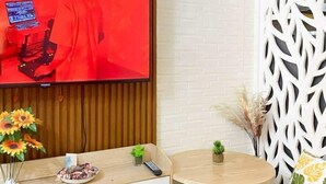 Uma sala de estar moderna com uma TV de tela plana fixada em uma parede de madeira, uma mesa de madeira redonda com duas cadeiras, uma pequena planta em vaso e um divisória branco decorativo com padrão geométrico.