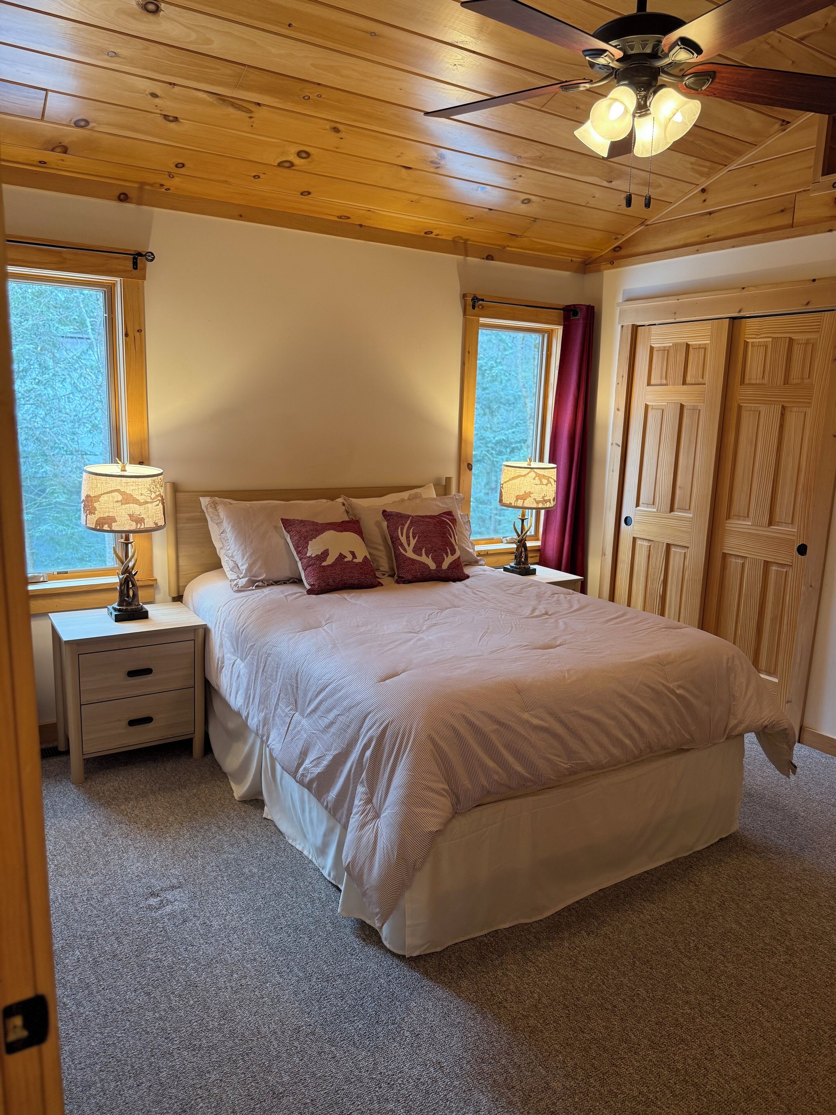 A bedroom with a wooden ceiling, a bed with a beige comforter, two bedside tables with lamps, and a window with a view of trees.