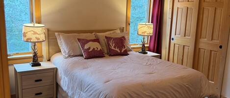 A bedroom with a wooden ceiling, a bed with a beige comforter, two bedside tables with lamps, and a window with a view of trees.