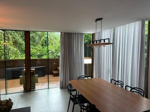 Uma sala de jantar moderna com mesa e cadeiras de madeira, grandes janelas com cortinas e vista para um deck de madeira e vegetação lá fora.