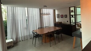 Un espace repas moderne avec une table et des chaises en bois, de grandes fenêtres avec des rideaux et, au fond, un coin cuisine.