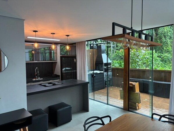 Uma cozinha moderna com uma ilha central, uma sala de jantar com mesa de madeira e uma porta de vidro deslizante que dá para um deck com churrasqueira.