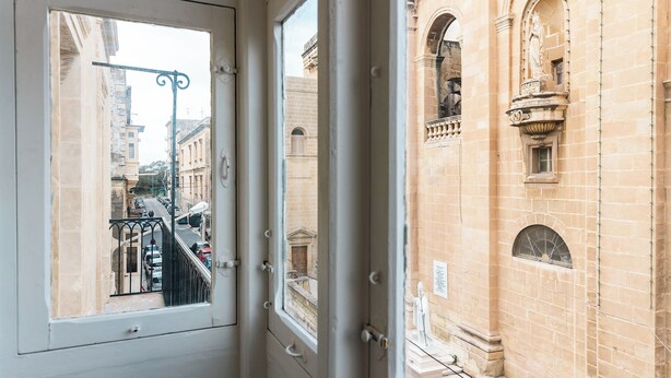 A view through a window of a building with a balcony, a street, and a statue. - Florette Suites (Floriana)
