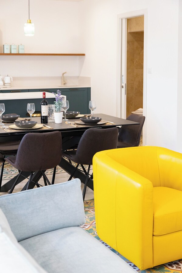 A dining area with a table set for four, chairs, a yellow armchair, and a shelf with kitchen items.