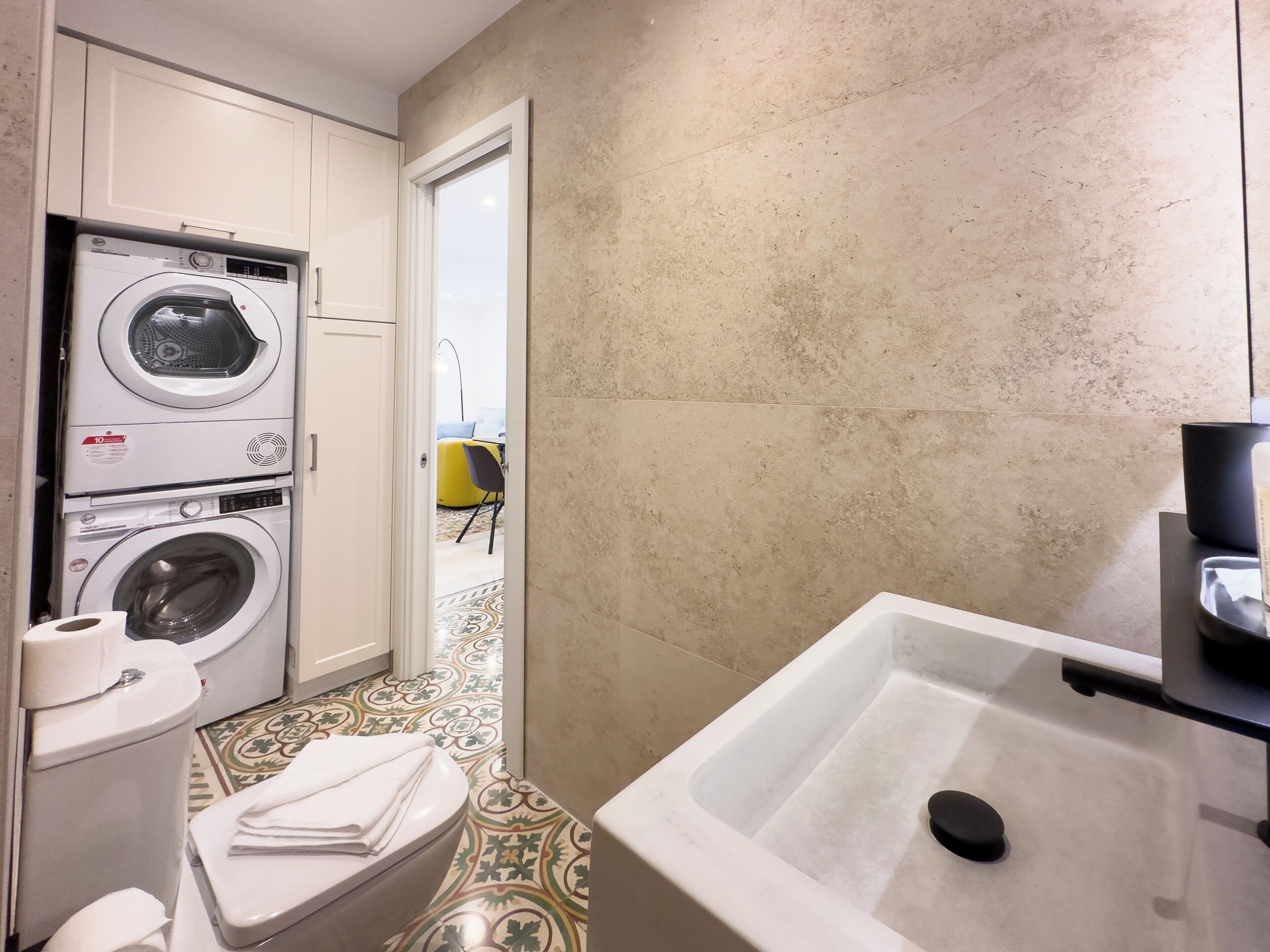 A compact hotel bathroom with a washing machine, toilet, sink, and patterned floor tiles.