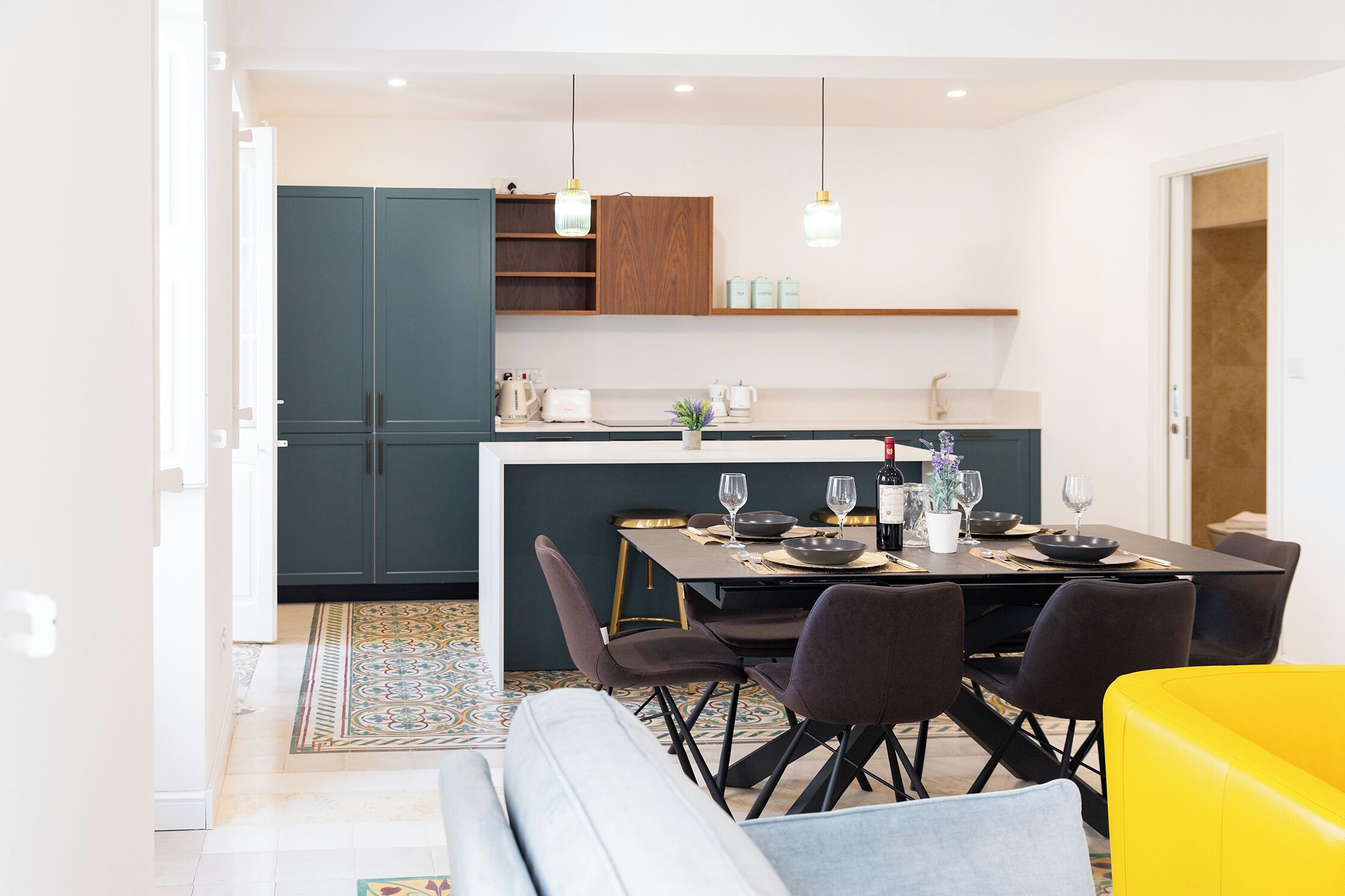 A modern kitchen with dark cabinets, a dining table set for four, and a yellow sofa in the foreground.