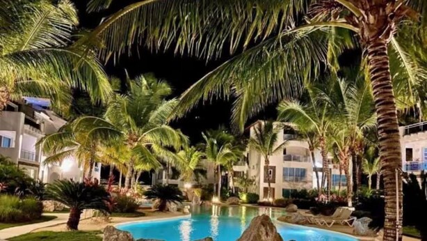 Vista nocturna de una piscina rodeada de palmeras y un edificio con varios balcones.