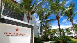 Entrada de un hotel con un cartel que dice "ESTRELLA DOMINICUS" y un camino que conduce al edificio, rodeado de palmeras.