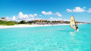 Una playa con agua turquesa cristalina, una persona practicando windsurf y un complejo turístico con edificios al fondo.