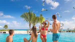 Escena junto a la piscina con gente relajándose, palmeras y un cielo azul despejado.