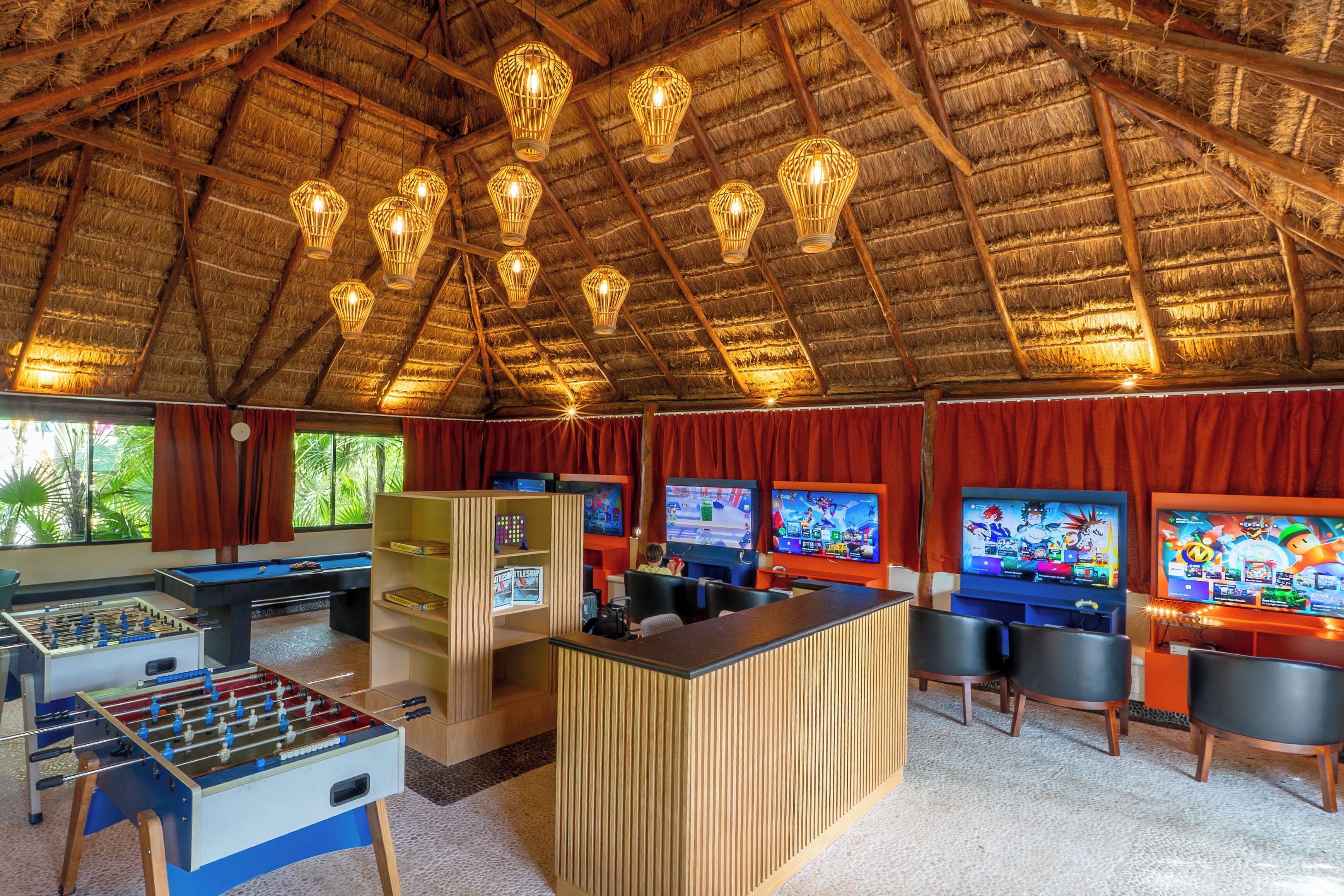 Een kamer met rieten dak, houten balken en een centrale ruimte met een tafelvoetbaltafel, twee spelconsoles en een tafelvoetbaltafel.