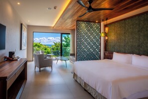 Habitación de hotel moderna con una cama grande, un escritorio con cafetera, una silla y un balcón con vistas a la vegetación y al cielo.