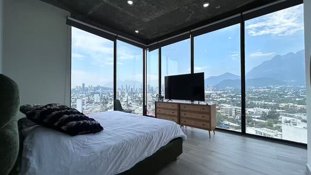Un dormitorio con una cama grande, un televisor en una cómoda y vistas a la ciudad a través de amplias ventanas.
